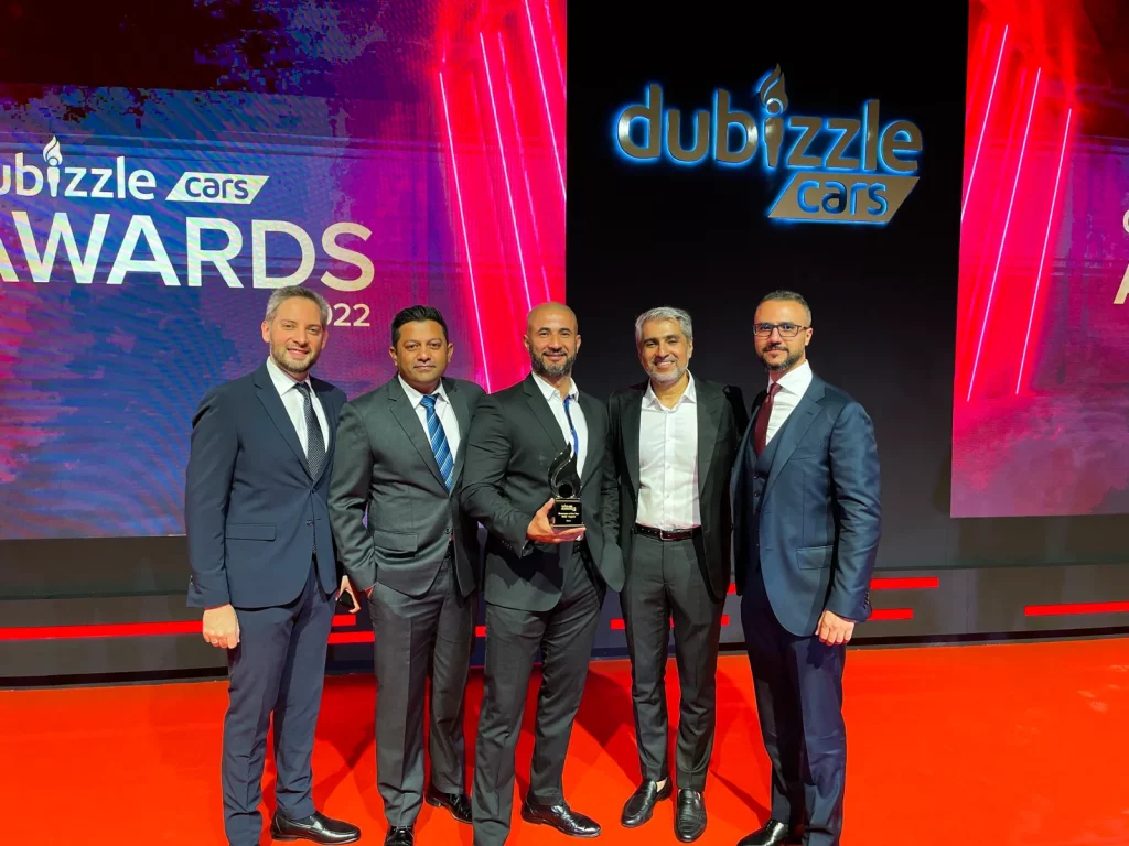 Five GTA Cars team members in suits on stage at the Dubizzle Cars Awards 2022, one holding the trophy, in front of the illuminated dubizzle cars backdrop