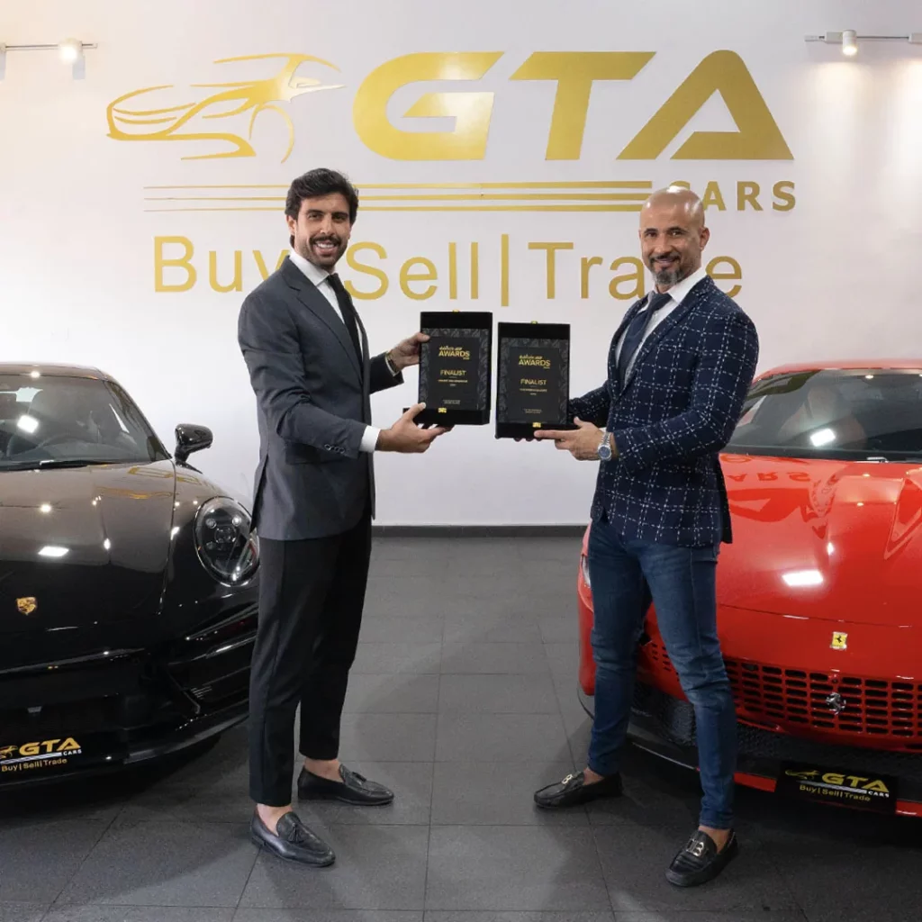 Two GTA Cars team members holding a pair of Dubizzle Cars Awards finalist plaques in the showroom, standing between a black Porsche 911 and a red Ferrari beneath the GTA Cars wall logo