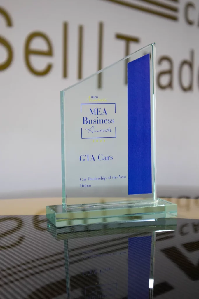 MEA Business Awards glass trophy engraved “GTA Cars — Car Dealership of the Year, Dubai”, standing on a desk in front of the GTA Cars Buy | Sell | Trade wall sign