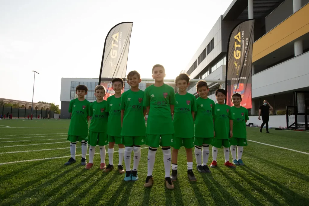 Young Football Academy players in green kit lined up on a grass pitch between GTA Cars branded flags, marking GTA Cars' 2021 sponsorship of The Football Academy.